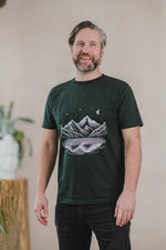 A man models a green t-shirt with a mountain and star design, showcasing the Moonlit Peaks Men's Organic T-shirts