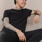 A Tattooed Man Sitting On A Couch, Wearing A Stylish Organic T-shirt, Relaxed And Casual In His Environment.