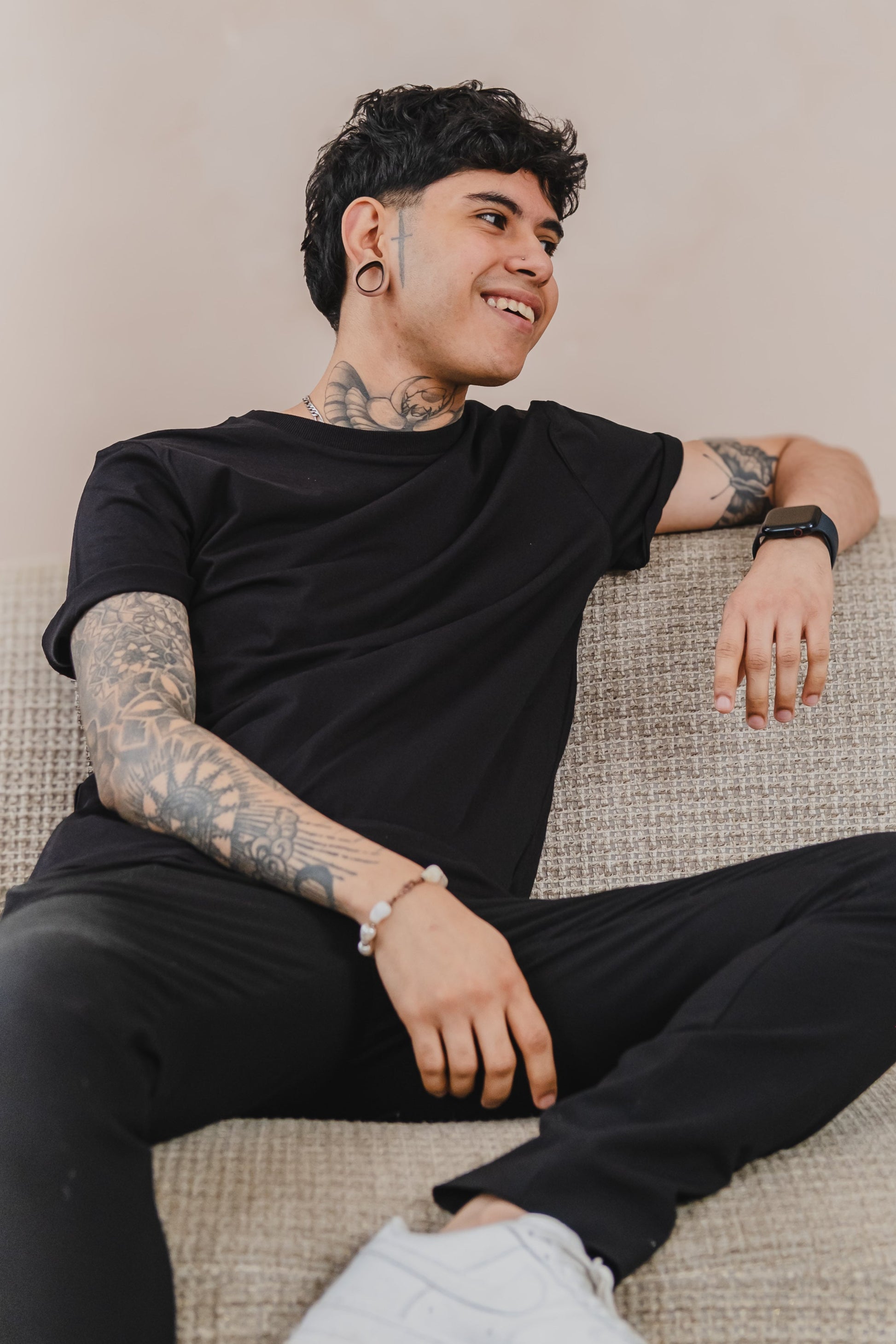 A Tattooed Man Sitting On A Couch, Wearing A Stylish Organic T-shirt, Relaxed And Casual In His Environment.