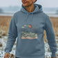A Woman In A Blue Hoodie Featuring A Bird And Wave Design, Showcasing The Sun And Birds Women's Pullover Organic Hoodie.