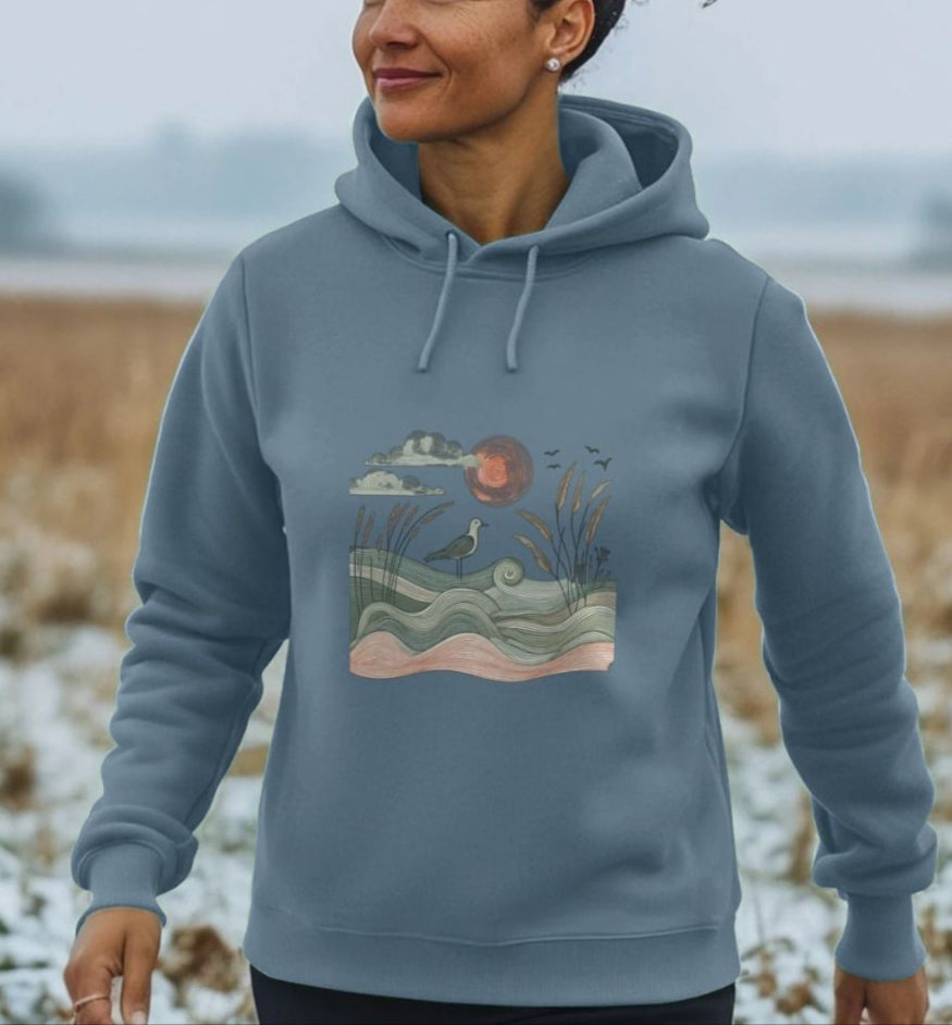 A Woman In A Blue Hoodie Featuring A Bird And Wave Design, Showcasing The Sun And Birds Women's Pullover Organic Hoodie.