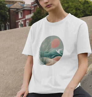 A woman on a bench wears a white t-shirt with a bird and sunset design